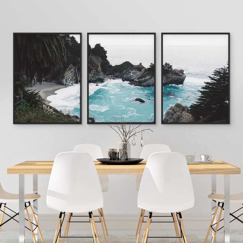 May include: Three framed photographs of a rocky coastline with turquoise water and lush green trees. The photos are hung on a white wall above a wooden table with white chairs.