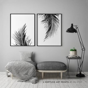 May include: Two black and white prints of palm leaves, framed in black, hanging on a grey wall above a grey ottoman and a black side table with a potted plant.