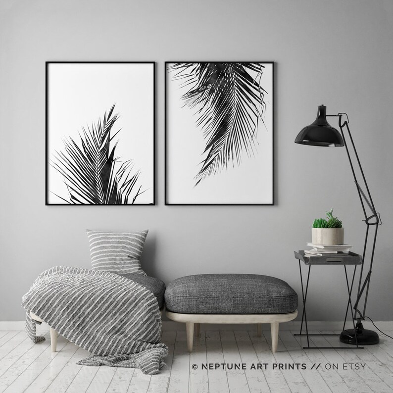 May include: Two black and white prints of palm leaves, framed in black, hanging on a grey wall above a grey ottoman and a black side table with a potted plant.
