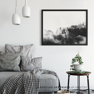 May include: Black and white photograph of a foggy forest scene. The image is framed in a black frame and hangs on a white wall above a gray couch with pillows and a throw blanket.