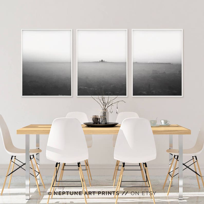 May include: Three black and white landscape photographs of a foggy scene with a small building in the distance. The photographs are framed in white and hung on a white wall above a wooden table with white chairs.