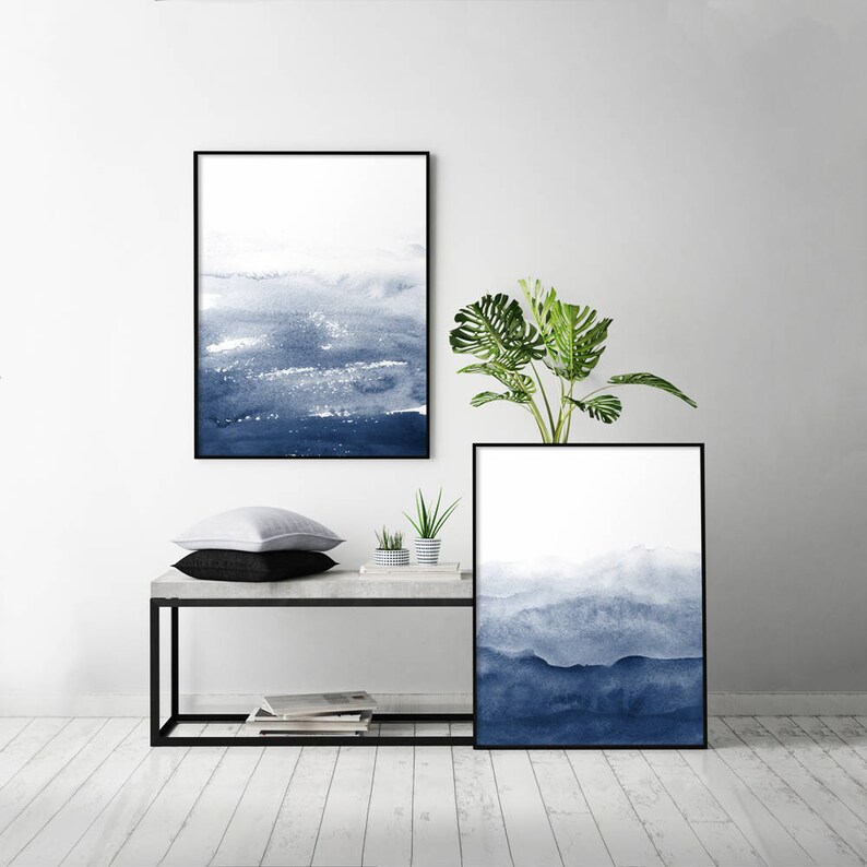 May include: Two framed watercolour paintings with a blue and white colour scheme. The paintings are hanging on a white wall above a black metal shelf with a concrete top. A green plant is on the shelf.
