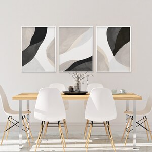 May include: Three abstract art prints in black and white with beige accents hang on a white wall above a wooden dining table with white chairs.
