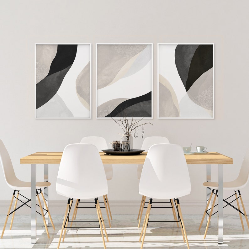 May include: Three abstract art prints in black and white with beige accents hang on a white wall above a wooden dining table with white chairs.