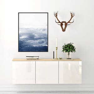May include: A white cabinet with three doors, a wooden top, and a blue and white abstract painting hanging above it. A deer antler mount is on the wall to the right of the painting.