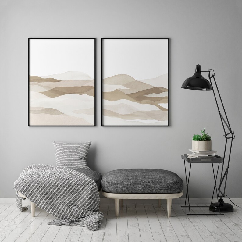 May include: Two framed abstract watercolor prints with brown and beige layered hills on a white background. The prints are hanging on a light gray wall above a gray ottoman with a white and gray striped throw blanket. A black floor lamp with a black shade is in the corner of the room next to a black side table with a potted plant.