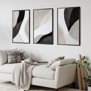 May include: Three abstract art prints with black and white geometric shapes on a white background. The prints are framed in black and hung on a white wall above a white couch with a throw blanket.