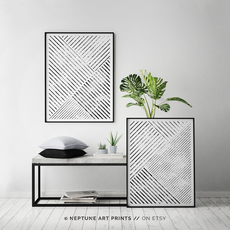May include: Two black framed prints with a white background and black diagonal lines. The prints are hanging on a white wall above a concrete shelf with a black metal frame. There are two small potted plants on the shelf and a white pillow on top of a black pillow.