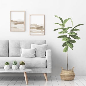 May include: A white sofa with two throw pillows and three potted succulents sits in front of a white wall with two framed abstract landscape prints. A large potted plant with green leaves stands to the right of the sofa.