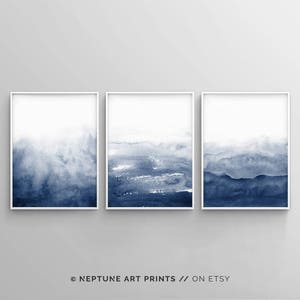 May include: Set of three abstract watercolour prints in shades of blue. The prints are framed in white frames and feature a gradient of blue from light to dark.