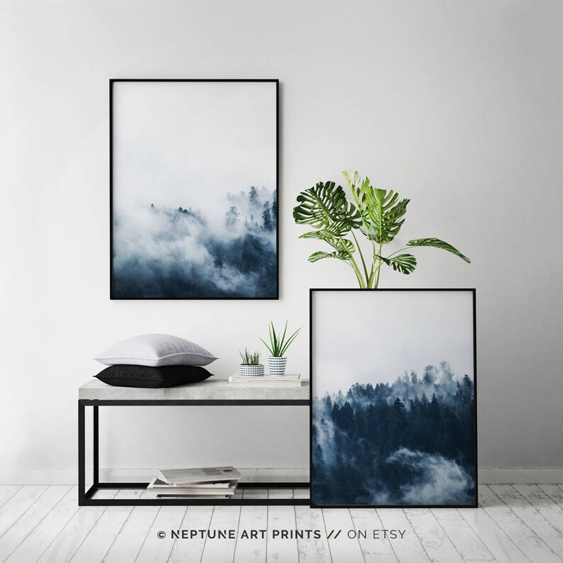 May include: Two framed prints of a misty forest scene in shades of blue and white. The prints are hanging on a white wall above a concrete shelf with a black metal frame. A potted plant with green leaves is on the shelf.