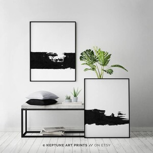 May include: Two black and white abstract art prints with a black brushstroke design. The prints are framed in black and are hanging on a white wall. A green plant is in front of the prints.