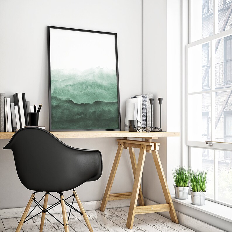 May include: A black framed print of a watercolour painting of a green mountain landscape hangs above a wooden desk with a black chair and a wooden easel. Two small potted plants are on the windowsill.