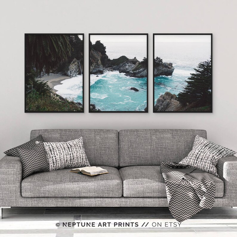 May include: Three framed photographs of a rocky coastline with turquoise water. The photos are arranged in a row on a gray wall above a gray couch with pillows and a throw blanket.