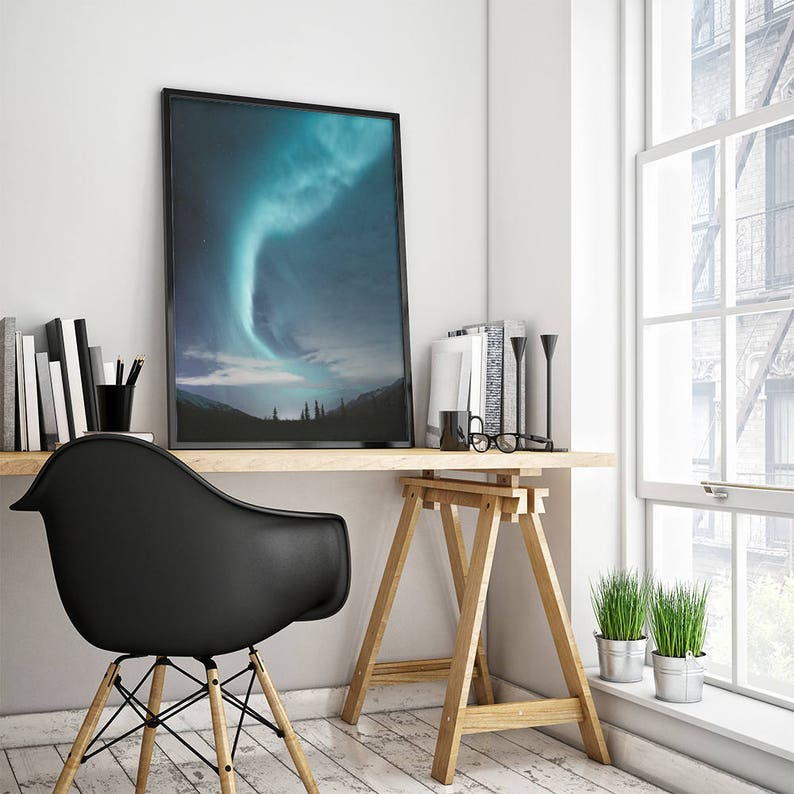 Puede incluir: Una impresi&oacute;n enmarcada en negro de una aurora boreal verde y azul sobre un bosque oscuro. La impresi&oacute;n est&aacute; colgada en una pared blanca sobre un escritorio de madera con una silla negra, un caballete de madera y dos plantas en maceta.