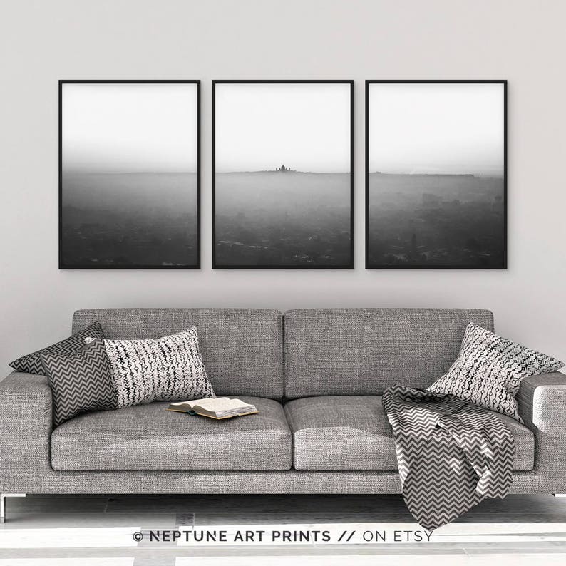 May include: Three black and white photographs of a foggy landscape with a silhouette of a city in the distance. The photographs are framed in black frames and are hung on a white wall above a gray couch.