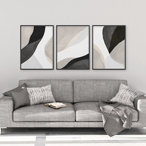 May include: Three abstract art prints in black frames, each featuring a different color scheme of black, white, and beige. The prints are hung on a white wall above a gray couch with pillows and a throw blanket.