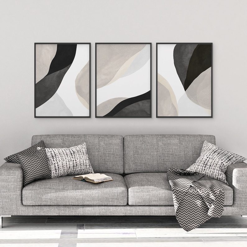 May include: Three abstract art prints in black frames, each featuring a different color scheme of black, white, and beige. The prints are hung on a white wall above a gray couch with pillows and a throw blanket.