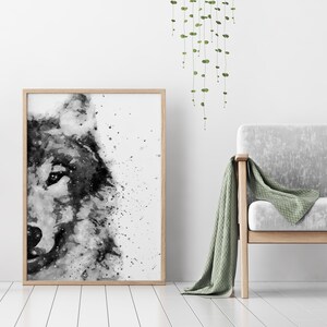 May include: A black and white watercolour painting of a wolf's head, framed in a light wood frame. The painting is hanging on a white wall with a green plant hanging to the right.