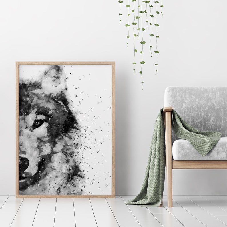 May include: A black and white watercolour painting of a wolf's head, framed in a light wood frame. The painting is hanging on a white wall with a green plant hanging to the right.