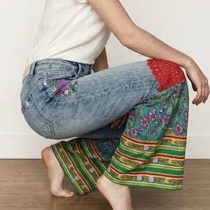 May include: Light blue denim jeans with floral embroidery and patterned fabric inserts. The jeans feature a red crocheted knee patch. A white t-shirt is also visible. The model is squatting.
