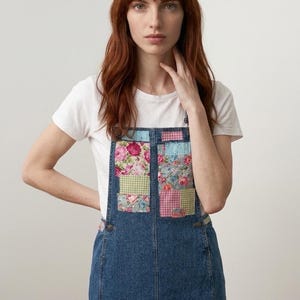 May include: A denim dungaree dress with floral patchwork pockets and a heart-shaped patch. The dress has a classic bib design with adjustable straps and a knee-length skirt. The patchwork includes pink, blue, and green floral and gingham patterns.