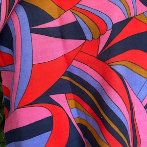 May include: Abstract fabric with a geometric pattern in shades of red, pink, blue, and brown. The fabric has a woven texture.