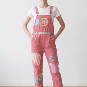 May include: A pair of pink corduroy dungarees worn over a white t-shirt. The dungarees are embellished with assorted fabric patches, including heart shapes, a balloon motif, a circular crochet doily on the chest, and white trainers.