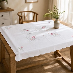 May include: A white square tablecloth with scalloped edges, featuring delicate floral embroidery of pink roses and green leaves arranged in a central square pattern and at each corner, covering a wooden table surface.