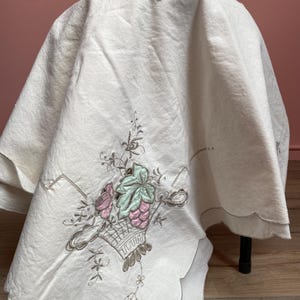 May include: Off-white tablecloth with a scalloped edge. The tablecloth features an embroidered design of a basket filled with grapes and flowers in shades of pink, green, and brown. The tablecloth is draped over a table.