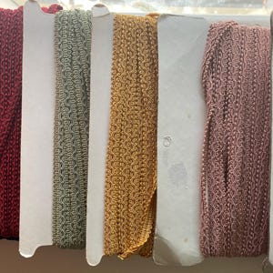 May include: A selection of decorative trim in various colours, including navy, burgundy, olive green, gold, rose, and white. The trim has a textured, woven design and is displayed on cardboard spools. Ideal for sewing and craft projects.