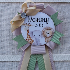 May include: A green, gold, and pink ribbon rosette with a round button featuring a giraffe, elephant, and lion with the text "Mommy to Be".