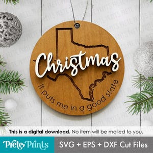 May include: A round wooden Christmas ornament featuring the state of Texas. The word "Christmas" is in white script across the state outline. The phrase "It puts me in a good state" is inscribed around the edge.