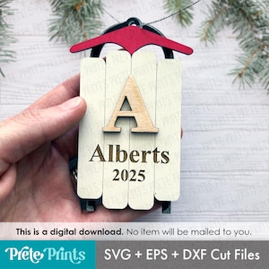 May include: A wooden Christmas ornament shaped like a sled. The sled is cream-colored with a red top and black runners. It features the letter "A", the name "Alberts", and the year "2025". The ornament is held by a hand.