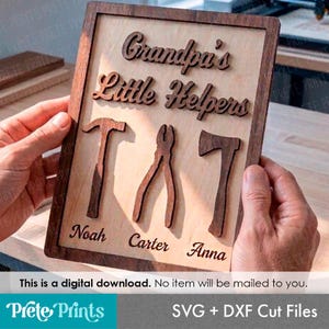 May include: A rectangular wooden sign featuring the laser-cut text Grandpa's Little Helpers in dark wood against a light wood background. Below the text are three wooden tool cutouts: a hammer, pliers, and an axe, with the names Noah, Carter, and Anna.