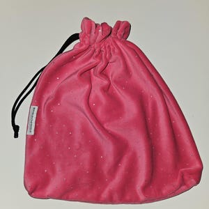 May include: A pink velvet drawstring bag with a sparkling finish. The bag has a gathered top and a black drawstring closure. The bag is made of a soft, plush material and is designed to hold small items.