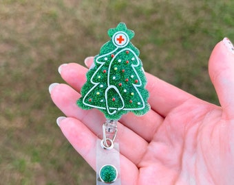 May include: A green glitter Christmas tree with a red cross on top, attached to a clear plastic retractable badge holder with a green glitter button.