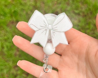 May include: White glitter bow badge reel with a white pearl charm and a clear plastic badge holder.