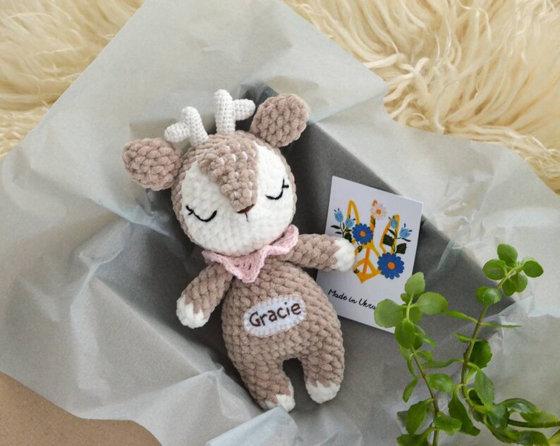 以下が含まれることがあります： 茶色、白、ピンクの色合いの手作りの編みぐるみ鹿のぬいぐるみ。目は閉じられ、ピンク色の襟が付いています。「Gracie」という名前が腹部に刺繍されています。「Made in Ukraine」と書かれた花の模様のカードも表示されています。