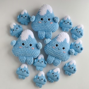 May include: A collection of crocheted mountain plushies in various sizes. The mountains are light blue with white snowcaps and smiling faces. The plushies have small arms and legs, creating a whimsical design.