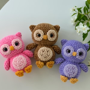 May include: Three crocheted owl plushies in pink, brown, and purple. Each owl features large dark eyes, a small yellow beak, a round belly patch, and two small yellow feet. The plushies are crafted from soft yarn with a textured, looped appearance.