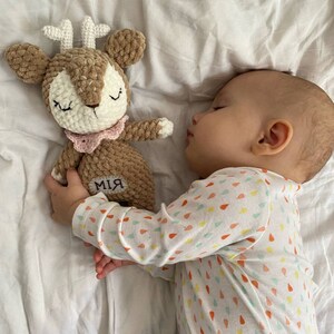 以下が含まれることがあります： 茶色、クリーム色、白のグラデーションの、かぎ針編みの鹿のぬいぐるみ。ピンクの襟と体に「MIA」と刺繍されています。鹿は目を閉じており、模様入りのロンパースを着た赤ちゃんの隣に横たわっています。