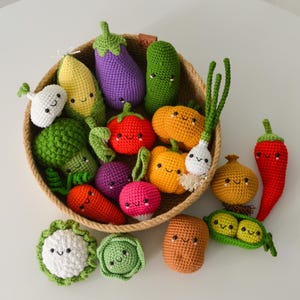 Könnte beinhalten: Eine Sammlung von gehäkelten Gemüse-Spielzeugen mit gestickten lächelnden Gesichtern, darunter Mais, Aubergine, Brokkoli, Tomate, Paprika, Zwiebel und Knoblauch, angeordnet in einem runden geflochtenen Korb mit weiteren losen Teilen daneben.