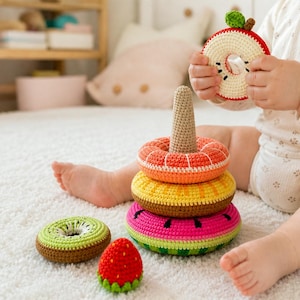 Puede incluir: Una colección de juguetes de ganchillo coloridos, que incluyen una torre de anillos apilables y piezas con forma de fruta. La torre presenta anillos naranjas, amarillos y rosas, coronados por un cono beige. Un niño sostiene un juguete con forma de manzana. Los juguetes están sobre una alfombra blanca.