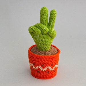 May include: A crocheted cactus shaped like a peace sign in a bright orange pot with white zig-zag pattern.