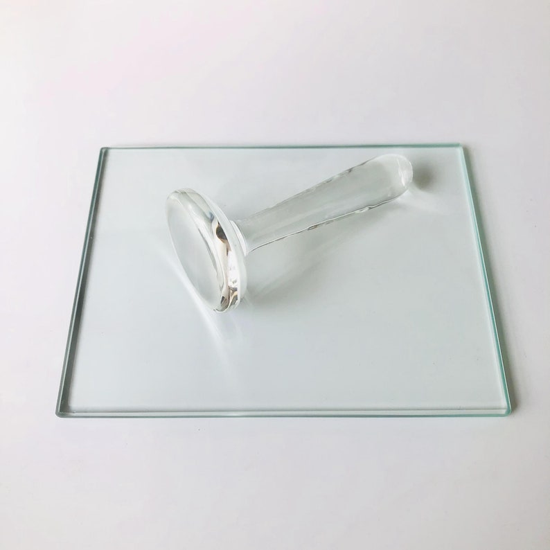 May include: A clear glass stamp with a round base and a long handle. The stamp is sitting on a clear glass square.