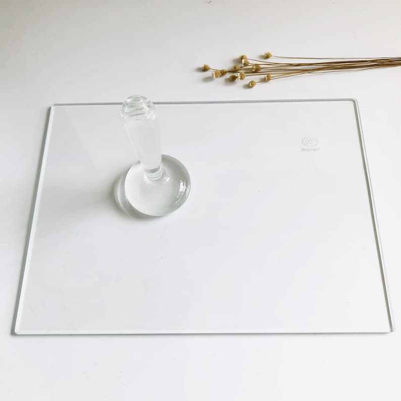May include: A clear glass cake stand with a round base and a tall, cylindrical pedestal. The stand is on a white surface.