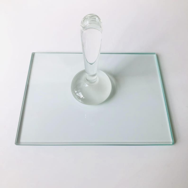 May include: A clear glass bottle stopper with a round base and a tall, slender neck. The stopper is sitting on a clear glass square plate.