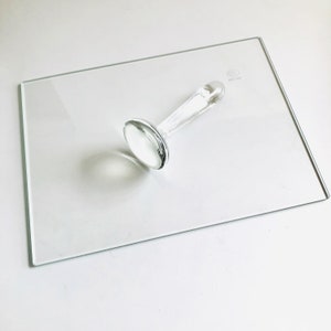 May include: A clear glass stamp with a round head and a long handle. The stamp is sitting on a clear glass plate.
