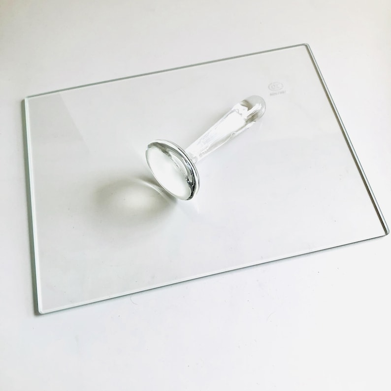 May include: A clear glass stamp with a round head and a long handle. The stamp is sitting on a clear glass plate.